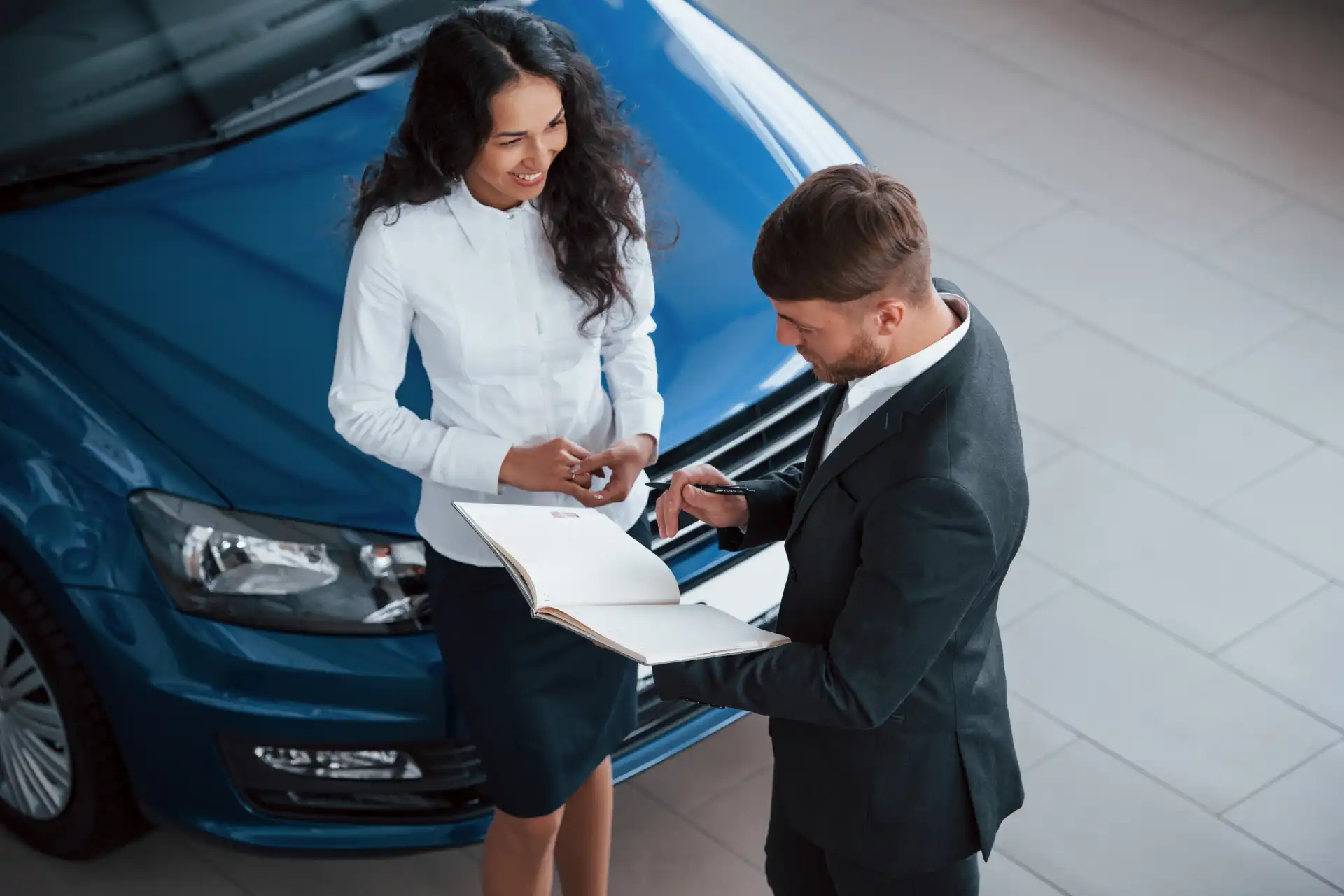 Homem e mulher conversando na concessionária de carros, com um carro azul ao fundo, enquanto olham um documento.