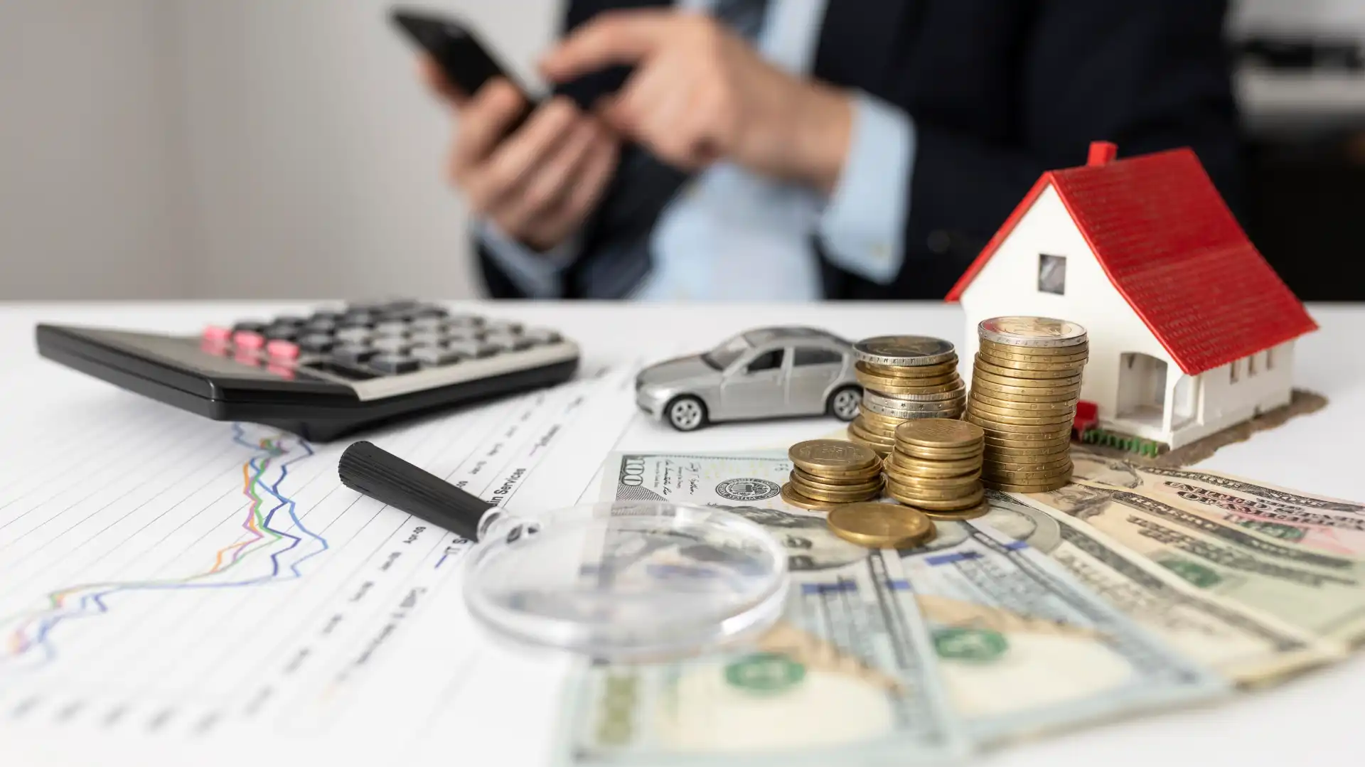 Imagem de plano de financiamento de imóveis com carro, moedas, casa em miniatura, gráfico e calculadora em uma mesa de escritório, representando investimento imobiliário.