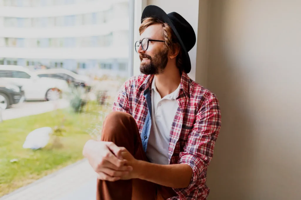 Seguros de vida para solteiros: Homem jovem com chapéu e óculos, sentado em um ambiente externo, com expressão pensativa, segurando o próprio joelho. Figura humana em contexto cotidiano.
